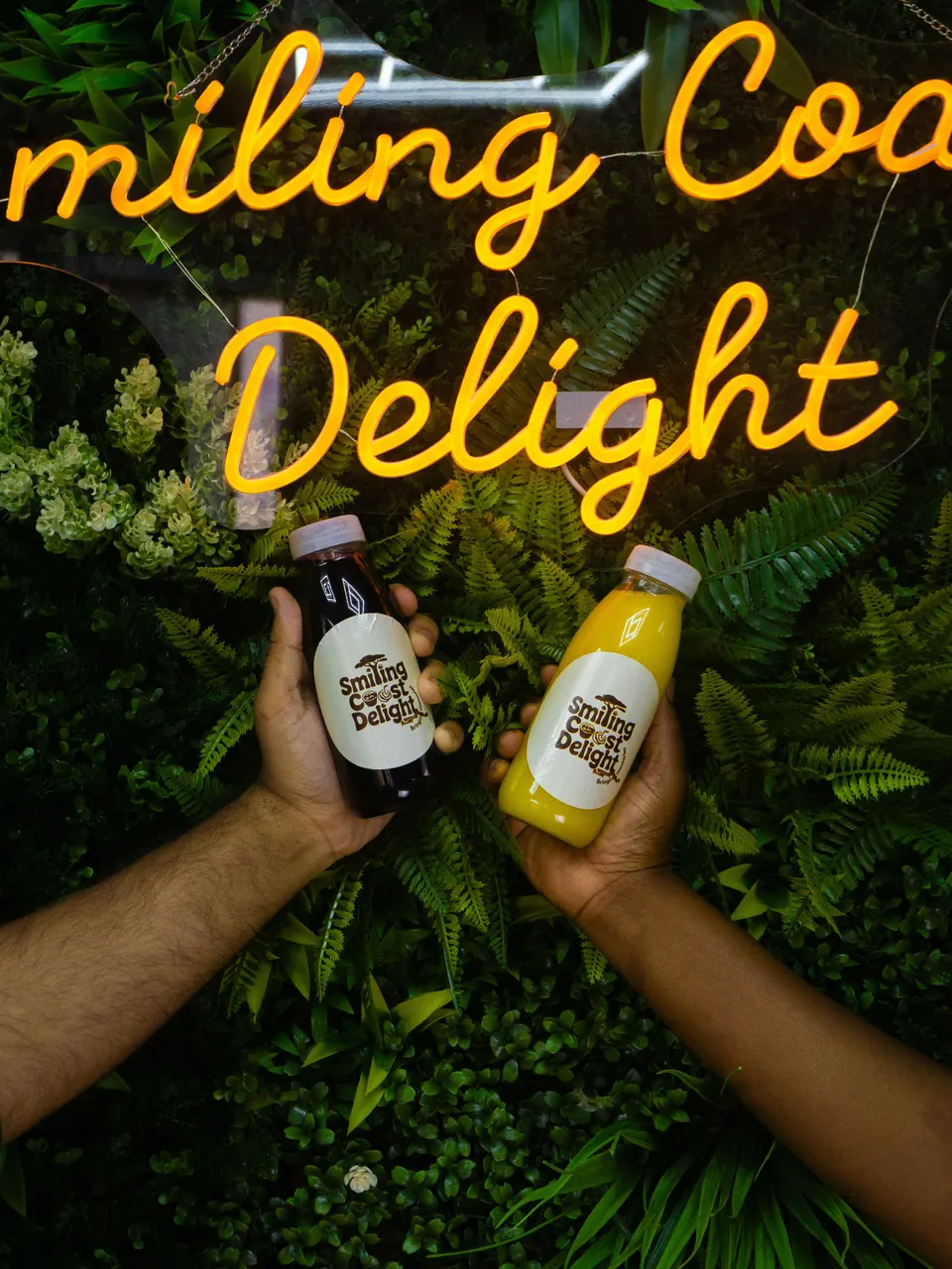 Two people holding bottled drinks beneath a neon Smiling Coast Delight sign